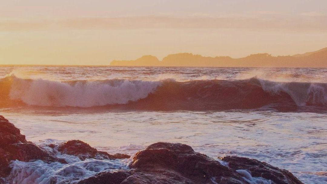 A wave breaking over dark rocks at sunset