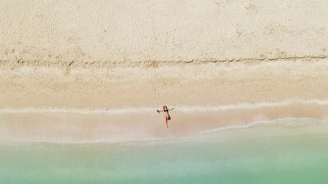 A person lying on a pale beach at the edge of clear green water, seen from above