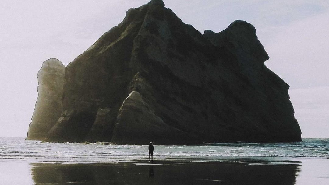 A lone figure standing below a huge rock rising out of still water