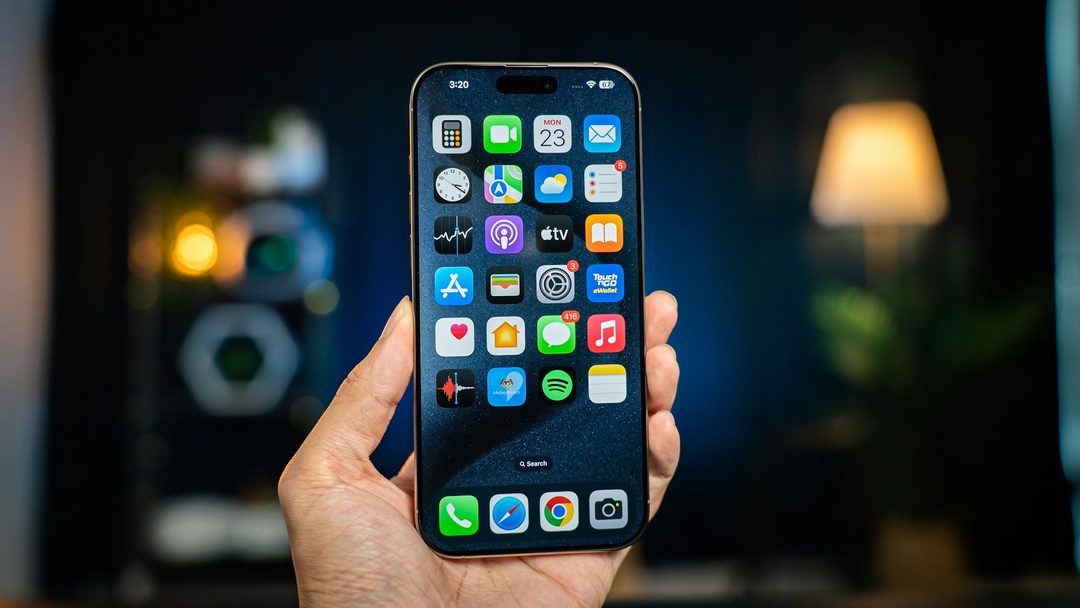 A hand holding an iPhone showing its Home Screen in a dim room
