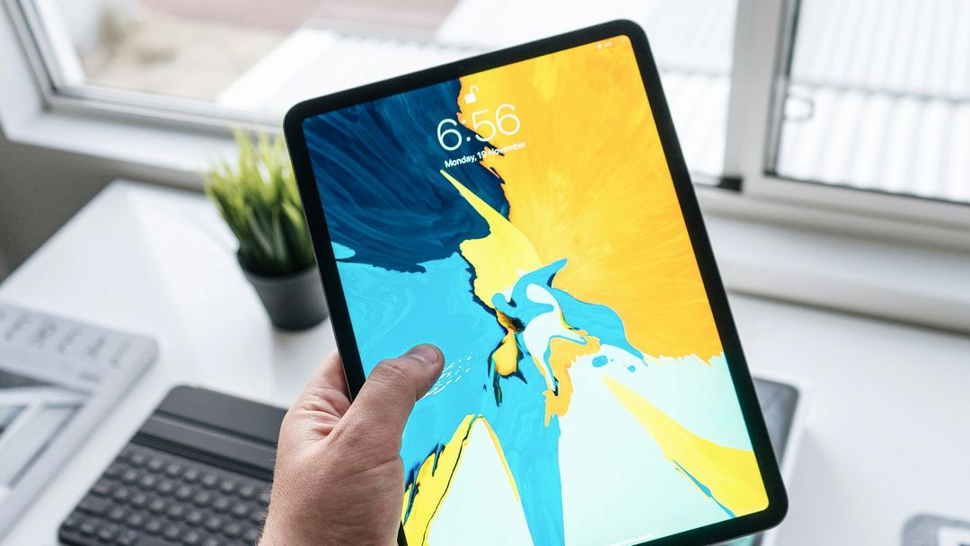 Uma mão segurando um iPad perto de uma janela clara, com a Tela Bloqueada mostrando a hora sobre uma imagem de fundo azul e amarela