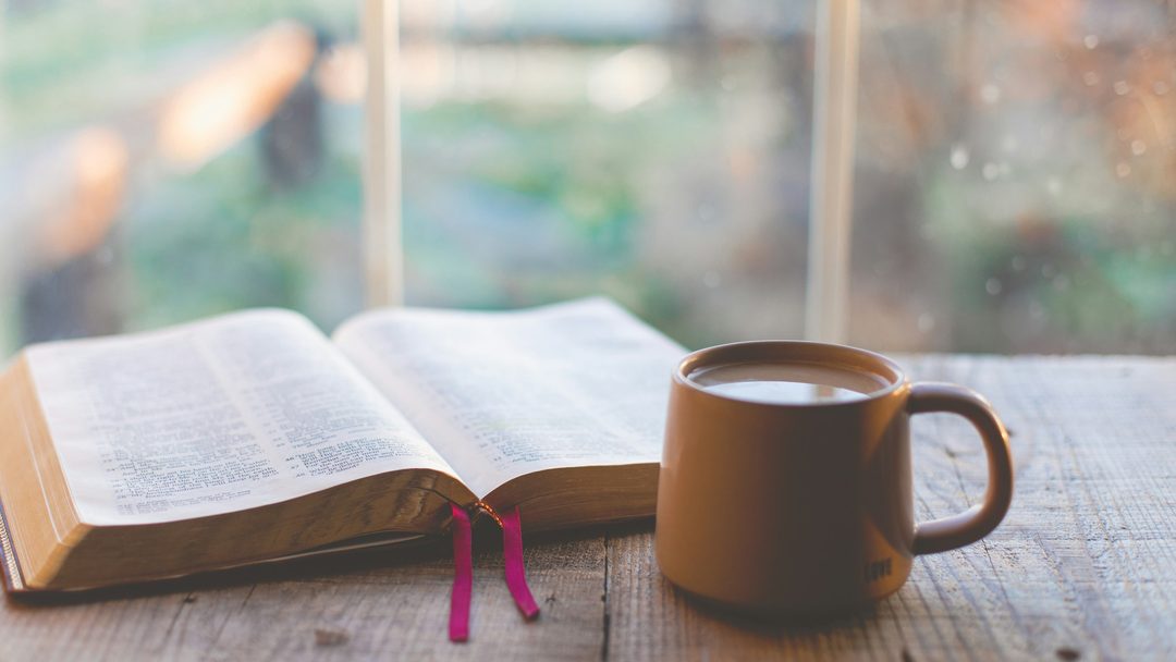 An open Bible beside a mug of coffee by a window