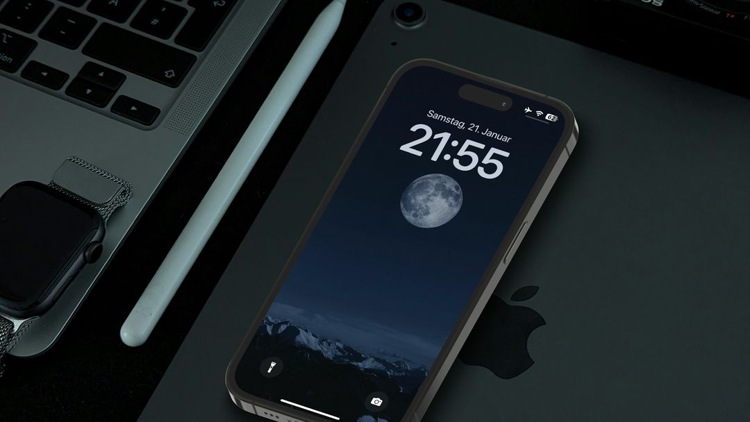 An iPhone on a dark desk beside a laptop and an Apple Watch, its Lock Screen showing the time over a full moon