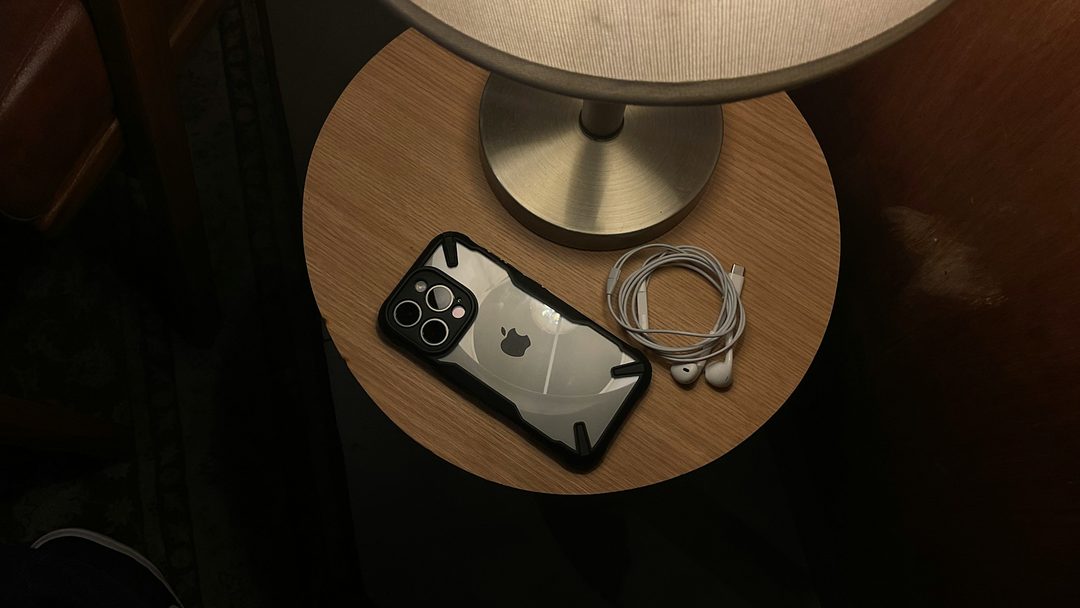 An iPhone and earbuds on a bedside table under a lamp
