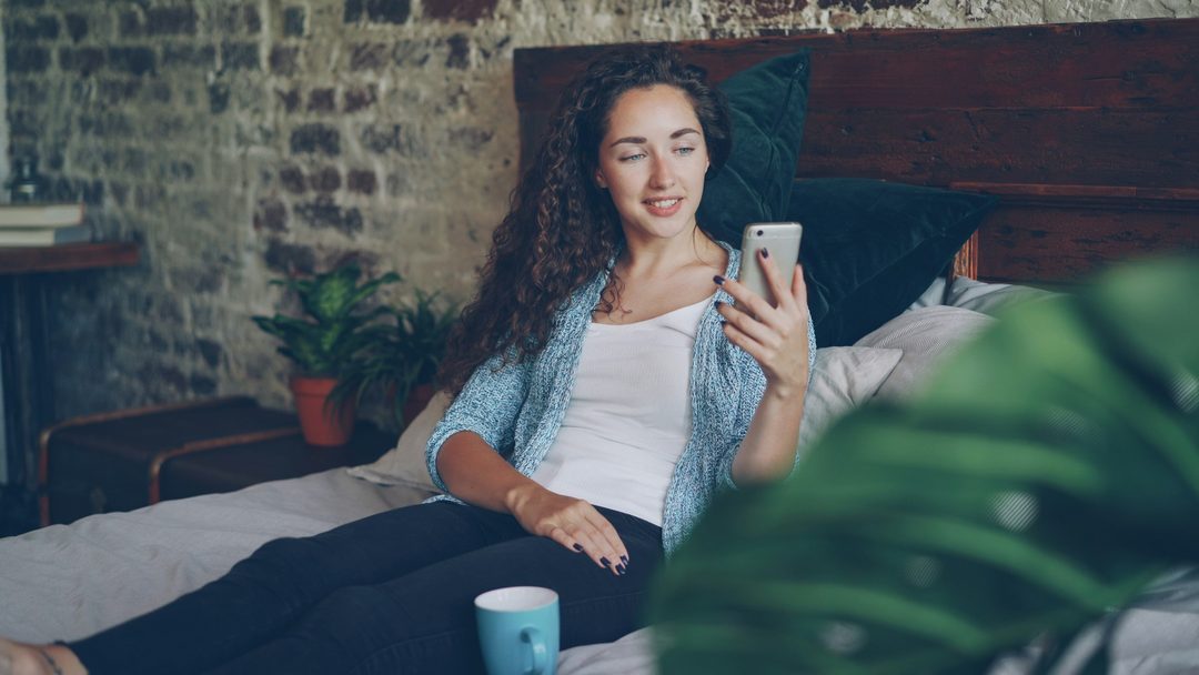 A woman relaxing in bed with her iPhone and a cup of coffee