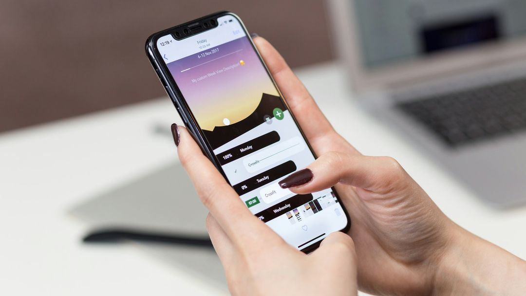 Two hands holding an iPhone over a white desk, with a laptop in the background
